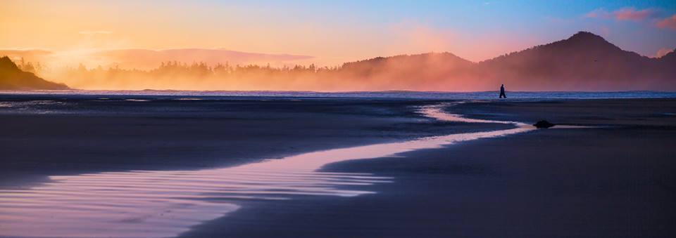 Person walking on a beach at sunset with mountains in the background.
