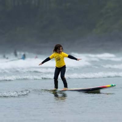 Child in yellow shirt surfing on small wave near coastline.