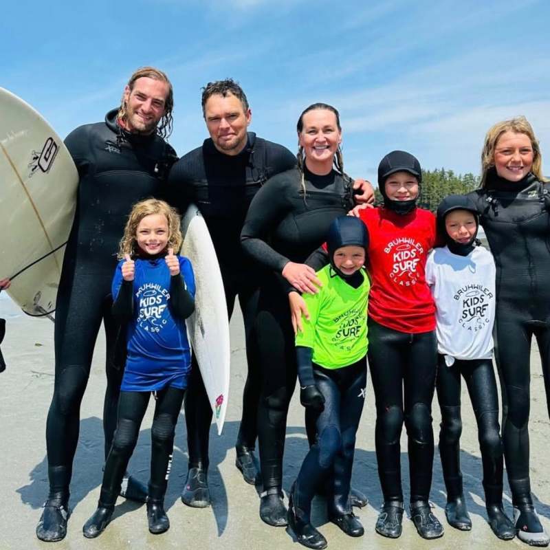 Group of adults and children in wetsuits holding surfboards on a beach.