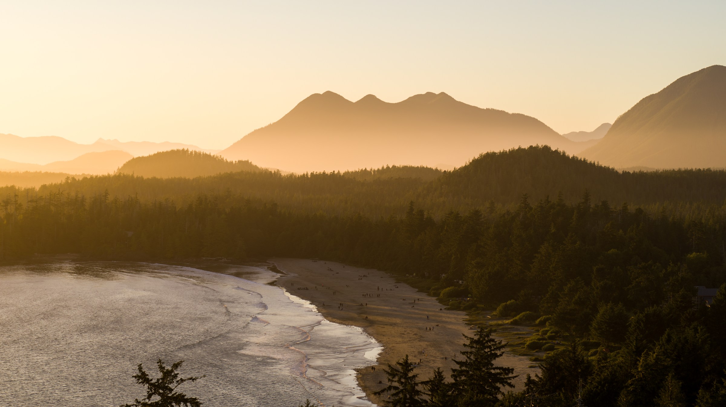 Sunset over a forested coastline with mountains in the background.