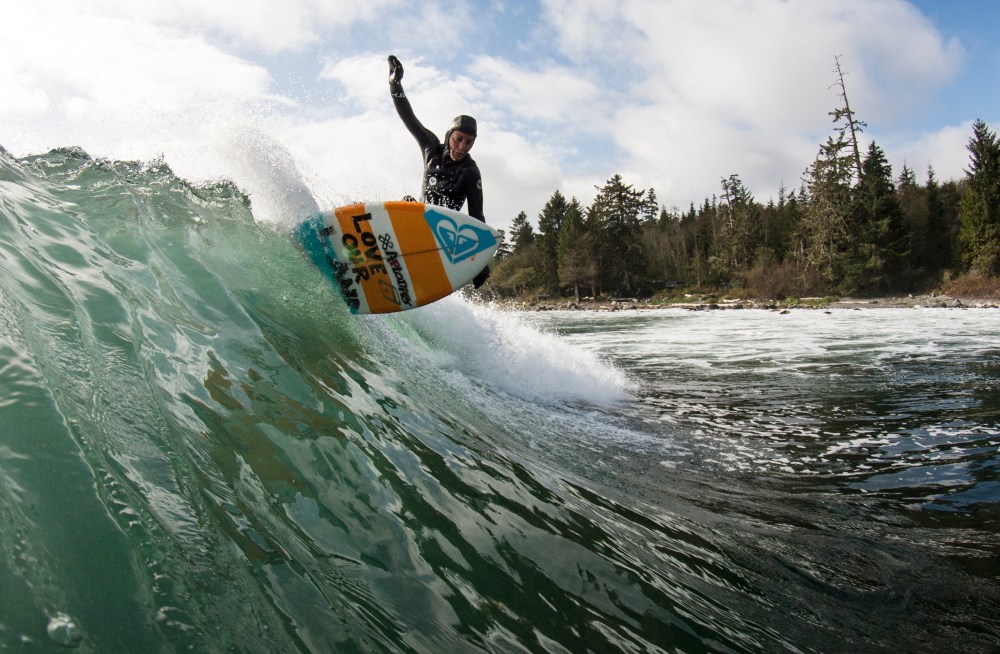 Surfer riding wave near forested shoreline under cloudy sky.